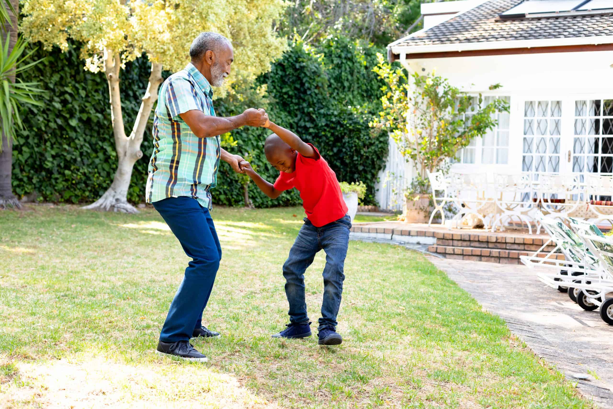 Senior man spending time with his grandson in their garden on a sunny day, teaching him dancing on the lawn.